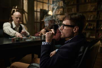 Stylish young man playing poker with his partners in a mysterious setting while smoking a cigar