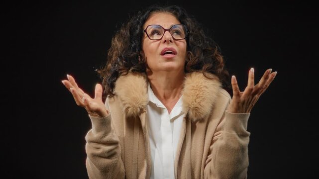Hispanic woman with curly hair and glasses gesturing expressively against an isolated black background wearing a fur-trimmed coat.
