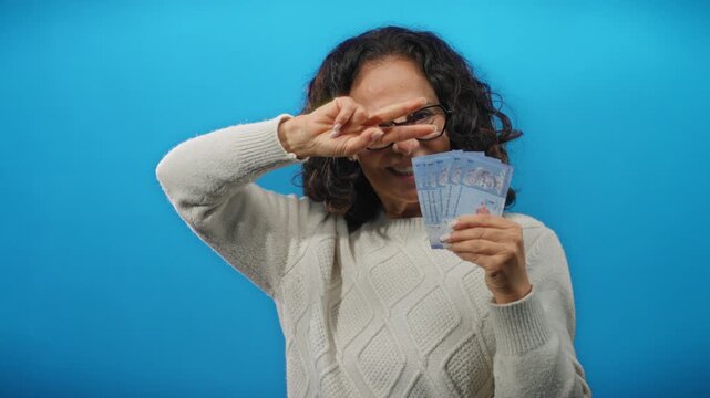 Middle-aged woman holding malaysian ringgit banknotes over blue background shows peace gesture with one hand in playful expression.