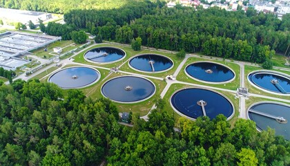 An aerial view of a wastewater treatment plant surrounded by lush greenery.  Circular basins and pathways are clearly visible within the facility's layout.