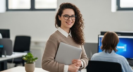 A smiling woman with curly hair and glasses holding a laptop in an office environment