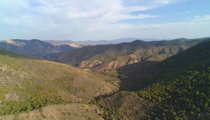 Fototapeta premium A high-angle view reveals a valley nestled between rolling hills, showcasing a mix of light brown and green vegetation, and a pale blue sky.