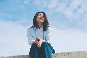 Cheerful young Asian woman sitting  on the roof under blue sky background.