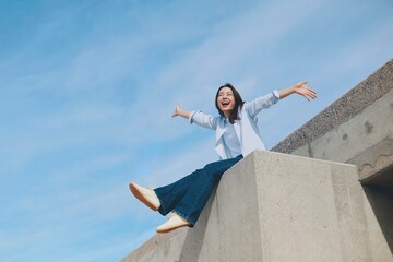 Cheerful young Asian woman sitting  on the roof under blue sky background.
