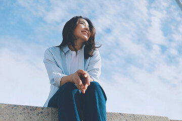 Cheerful young Asian woman sitting  on the roof under blue sky background.