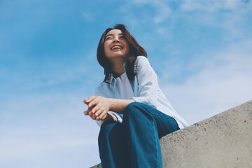 Cheerful young Asian woman sitting  on the roof under blue sky background.