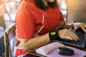 Obraz premium Hands of woman with thumb spica finger splint using computer mouse to work. Woman work on desk uses wrist support brace and using laptop computer. Broken hand in cast. Woman hands with orthosis.