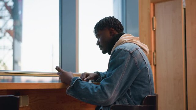 young black chap seated at wooden counter by window using smartphone while gazing at urban skyline through large pane from cafe interior with denim jacket hoodie casual modern vibe