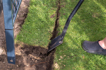 The photograph shows a man standing on a shovel with his foot. He is trying to stick the shovel into the lawn. Only the man's legs in dark clogs, the shovel and part of the lawn are visible.