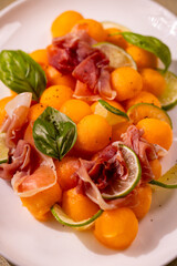 Melon and lime salad with prosciutto and basil leaves displayed on white plate