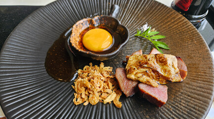 Foie gras with fried garlic and topped with cheese and yolk background on black dish. Sliced of beef or pork steak on brown plate.