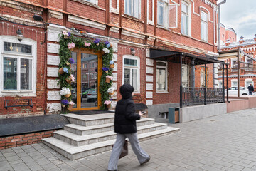 Historic brick building with a flower-decorated entrance and string lights. A pedestrian walks past on a revitalized city street.