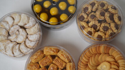 Five transparent plastic jars filled with: Nastar, Kastengel, Lidah Kucing, Putri Salju, and Chocolate Thumbprint Cookies. Each jar is neatly arranged in a row and isolated on a clean white background