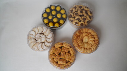 Five transparent plastic jars filled with: Nastar, Kastengel, Lidah Kucing, Putri Salju, and Chocolate Thumbprint Cookies. Each jar is neatly arranged in a row and isolated on a clean white background