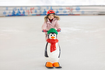 Happy little girl in warm pink coat and red safety helmet holding support and learning to skate on ice skating rink. Fall down and have fun. Winter sport, outdoors. Holiday and seasonal concept.