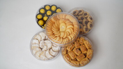 Five transparent plastic jars filled with: Nastar, Kastengel, Lidah Kucing, Putri Salju, and Chocolate Thumbprint Cookies. Each jar is neatly arranged in a row and isolated on a clean white background