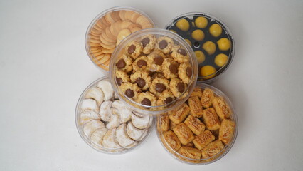 Five transparent plastic jars filled with: Nastar, Kastengel, Lidah Kucing, Putri Salju, and Chocolate Thumbprint Cookies. Each jar is neatly arranged in a row and isolated on a clean white background