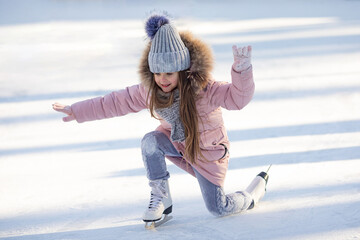 Cute little girl in warm pink coat, grey knitted hat and scarf, snood learning to skate on ice skating rink in park. Fall down and have fun. Winter sport, outdoors. Holiday and seasonal concept.