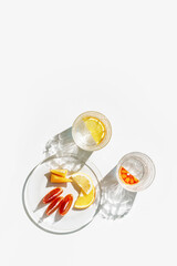 Summer still life scene with clear glass cups and plate. Glittering glasses of water with slices of fruit on white table at sunlight. Long harsh shadows and caustics. Summer vacation concept, above