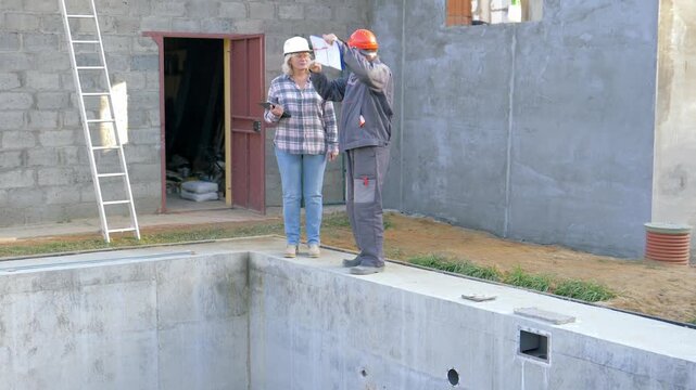 Two engineers a man and a woman, wearing protective helmet and holding blueprints and tablet, discuss plans next to an empty swimming pool under construction. Foreman and builder teamwork