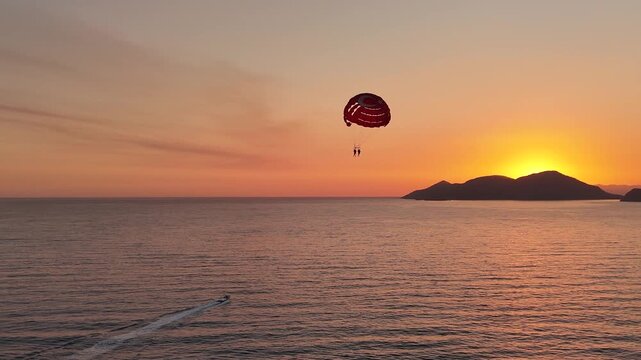 A breathtaking 4K aerial shot of two people parasailing in Oludeniz Fethiye their parachute with a Turkish flag silhouetted against a spectacular golden sunset over the calm sea