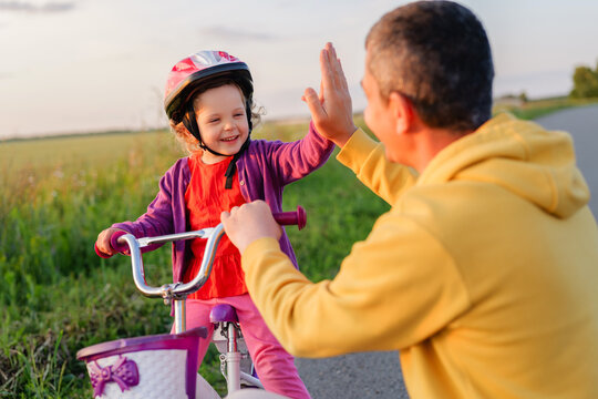 A smiling father gives his happy daughter a high five as she sits on her bike. She is wearing a helmet and a purple cardigan.