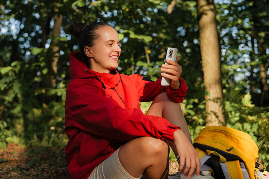 
A cheerful hiker in a red jacket smiles while looking at her phone, with a yellow backpack beside her in a forest.