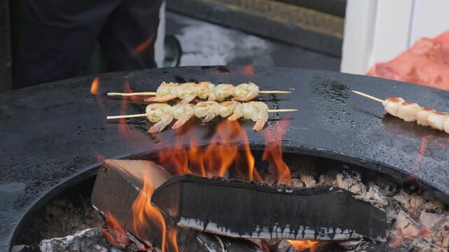 Street food chef in blue gloves grilling marinated shrimp skewers on circular grill above open flames during outdoor cooking at food market or festival