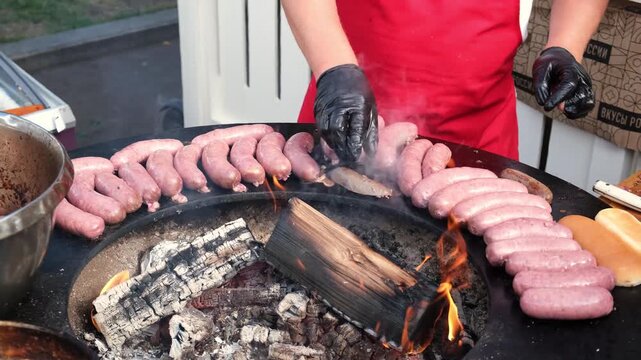 Street food vendor in red apron and gloves grilling fresh sausages over wood fire on a circular outdoor grill during food event or market