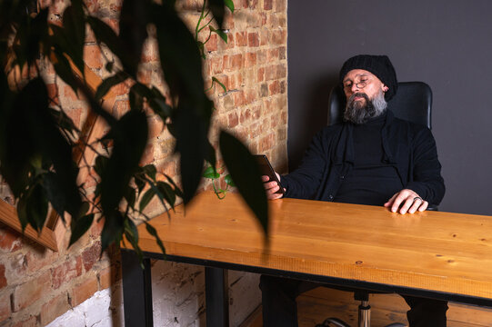 Portrait of a serious bearded hipster man in glasses and beanie sitting at a table with a smartphone. Creative freelancer in a loft office against a brick wall background