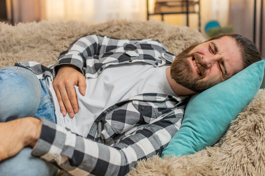 Young ill sick man lying at home with palm on abdomen, quietly moaning from stomach trouble or overeating. Caucasian guy on sofa curls slightly, enduring pain from potential gastritis or diarrhea.
