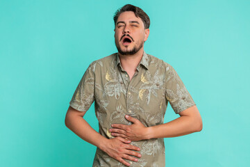 Middle-aged man holds stomach leans forward with tense expression indicating sudden pain or discomfort. Young adult guy isolated on blue background suffering from unexpected abdominal cramp or illness