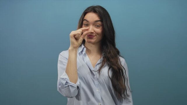Young hispanic woman pinching fingers to show small size while smiling in blue studio setting; playfulness.