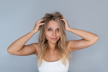 Fototapeta premium Young blonde woman with freshly dyed wavy hair touches her voluminous roots. Natural texture, porous hair type, and confident self-care moment on a light grey background