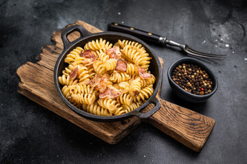 Fusilli carbonara pasta topped with crispy bacon bits and parmesan  cheese  in a skillet. Black background. Top view