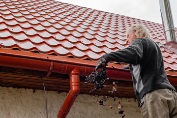 A man on a ladder clears leaves from the gutter of a house with a red tile roof