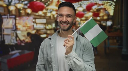 Hispanic man smiling and holding a nigerian flag in a vibrant urban street filled with lights and decorations, creating a lively multicultural atmosphere.