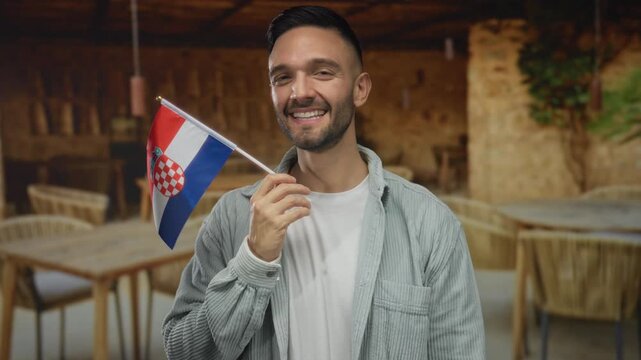Young man holding croatian flag smiling on restaurant terrace with tables in the background outdoors.