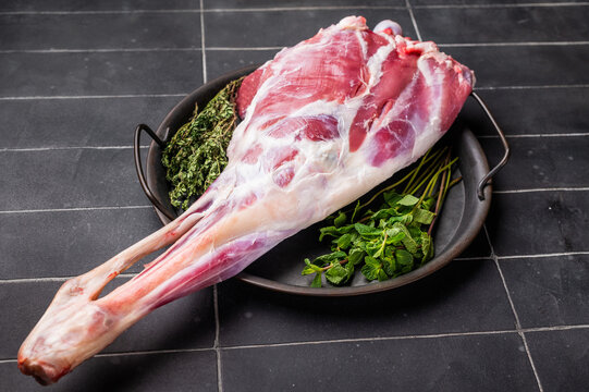 Uncooked whole goat leg, raw meat with herbs for cooking. black background. top view