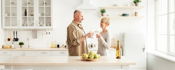 Happy couple in love have romantic dinner, toasting, enjoy home comfort and spare time. Cheerful adult female and man with glasses of white wine in modern kitchen interior, copy space, side view