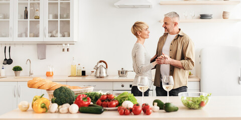 Proper nutrition, vegetarianism, romantic date during self-isolation at home. Happy senior male and lady dancing on modern kitchen interior with table with many fresh vegetables and glasses with wine