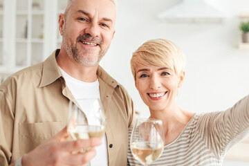 Video call congratulations, selfie at home and romantic dinner at covid. Portrait of smiling senior lady and male with glasses of white wine take photo in modern kitchen interior, close up, copy space