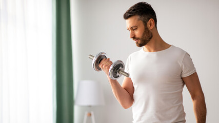 Closeup of handsome middle-aged man doing dumbbell workout at home, working on arms strength, looking at his biceps, copy space. Athletic man lifting dumbells up over living room interior