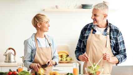Free time together, preparing lunch for family and having fun. Happy mature husband and wife make salad in kitchen interior with bright vegetables, glasses of wine and laptop on table, empty space