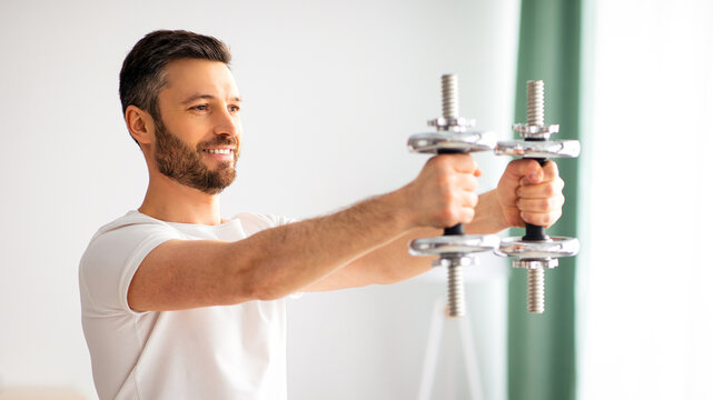 Athletic man lifting dumbells up over living room interior. Handsome middle-aged man doing dumbbell workout at home, working on arms strength, looking at copy space and smiling - Powered by Adobe