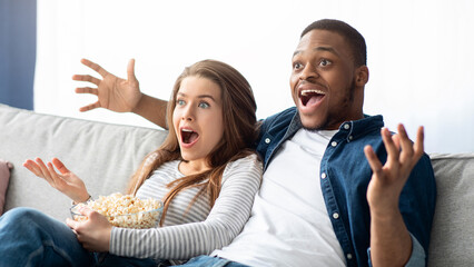 Excited interracial couple watching TV on couch at home, surprised black man and young woman emotionally reacting to interesting movie while relaxing in living room, sitting on sofa with popcorn
