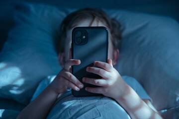 Close-up of unrecognisable little child lying in bed at night, holding mobile phone in front of his face. Gadget addiction and excessive late-night screen time. Smartphone overuse 