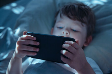 Fototapeta na wymiar Little child lying in bed in dark room, holding smartphone close and watching intently, illuminated by blue screen light. Gadget addiction and screen dependency. Selective focus on smartphone