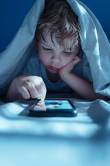 Little boy lying in bed under a blanket at night, intently watching and scrolling social media content on smartphone screen, face lit by blue light. Gadget addiction and overuse. Copy space