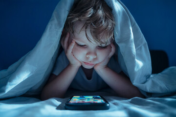 little bored boy in bed at night hiding under blanket, staring at glowing smartphone screen. Digital addiction, excessive screen time, and late-night gadget overuse.
